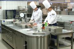 Akademia inspiracji Makro. Stanowisko kucharzy instruktorów. Kucharze instruktorzy przy pracy. obserwację ich działań ułatwiały ekrany z przekazem na żywo. Nagłowne zestawy mikrofonowe transmitowały instruktaż do sieci nagłośnieniowej sali szkoleń.Projekt stoiska i nadzór Tatjana Kobuszewska