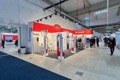 Stoisko targowe z czerwono-białym brandingiem, w tym logo "CAREL" i "ALFACO". Wzdłuż stoiska ustawione są panele i ekrany. Kilka osób przechodzi obok po wyłożonej kafelkami podłodze w dużej, jasnej hali wystawowej z wysokimi sufitami i górnym oświetleniem. projekt i nadzór Tatjana Kobuszewska trksystem.com