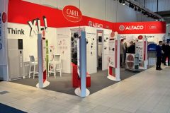 Stoisko targowe z czerwono-białym oznakowaniem dla CAREL i ALFACO. Na stoisku znajdują się wyświetlacze, plakaty, białe krzesła, stoły i kilku odwiedzających. Oświetlenie górne oświetla obszar, a podłoga jest wyłożona kafelkami w odcieniach bieli i beżu. projekt i nadzór Tatjana Kobuszewska trksystem.com