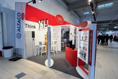 Stoisko targowe z czerwono-białym brandingiem CAREL prezentuje produkty i części HVAC. Stoisko zawiera slogan Think XL i ma półki z komponentami, ladę, krzesła i oznakowanie ALFACO. Hala wystawowa jest przestronna z podłogą wyłożoną kafelkami. projekt i nadzór Tatjana Kobuszewska trksystem.com