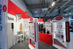 Stoisko targowe z czerwono-białym brandingiem Carel zawiera duże wyświetlacze produktów, panele informacyjne i wysokie stoły ze stołkami. Górne światła oświetlają eksponaty, a hasło "think XL" jest widoczne na zakrzywionej czerwonej ścianie z tyłu. projekt i nadzór Tatjana Kobuszewska trksystem.com