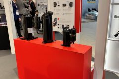 Jasnoczerwony stojak prezentuje cztery czarne sprężarki mechaniczne o różnych rozmiarach na stoisku targowym. Za nimi znajduje się plakat informacyjny marki Carel z tekstem i obrazami dotyczącymi technologii chłodniczej. W tle widoczni są ludzie i inne stoiska. projekt i nadzór Tatjana Kobuszewska trksystem.com