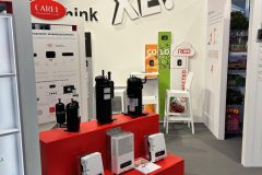 Stoisko targowe w odważnych czerwono-białych kolorach z napisem "XL" na ścianie. Sprężarki i nawilżacze marki CAREL są wyeksponowane na czerwonych półkach. Broszury, plakaty i białe krzesło są również widoczne na stoisku. projekt i nadzór Tatjana Kobuszewska trksystem.com