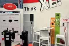 Stoisko targowe zawiera tekst Think XL na czerwono-białej ścianie, branding CAREL, plakaty informacyjne, wyświetlacze produktów z czarnymi kompresorami oraz część wypoczynkową z wysokim białym stołem i krzesłami. W tle znajdują się kolorowe banery produktowe. projekt i nadzór Tatjana Kobuszewska trksystem.com