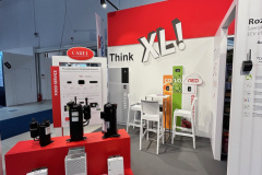 Stoisko targowe z czerwono-białym wystrojem prezentuje różne urządzenia przemysłowe na czerwonych stojakach. Na ścianie widnieje pogrubiony napis Think XL wykonany dużymi biało-czerwonymi literami. Mały stolik z krzesłami ustawiony jest pod ścianą obok kolorowych pionowych banerów. projekt i nadzór Tatjana Kobuszewska trksystem.com