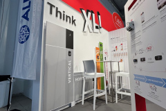 Stoisko targowe prezentuje sprzęt klimatyzacyjny lub HVAC. Na stoisku znajduje się duża biała pionowa jednostka z napisem VERTICAL obok wysokiego stołu z dwoma krzesłami. Czerwono-biały branding z Think XL i CAREL jest widoczny na ścianach. projekt i nadzór Tatjana Kobuszewska trksystem.com