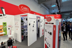 Stoisko targowe z czerwono-białym brandingiem CAREL, tablicami z informacjami technicznymi, próbkami produktów i kilkoma krzesłami. W tle widać kilka osób w strojach biznesowych, a górne światła oświetlają nowoczesną przestrzeń wewnętrzną. projekt i nadzór Tatjana Kobuszewska trksystem.com