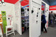 Stoisko targowe z białymi ścianami wyświetla informacje w języku polskim na temat rozwiązań ładowania pojazdów elektrycznych. Grafiki, wykresy i produkty są zamontowane na ścianach. Widoczne są dwie osoby; jedna stoi czytając, a druga jest częściowo zasłonięta za ścianą. projekt i nadzór Tatjana Kobuszewska trksystem.com