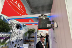 Mężczyzna w garniturze stoi na stoisku targowym pod zamontowanym urządzeniem wentylacyjnym. Stoisko ma białe ściany, stołki, stół i stojaki z roślinami. Czerwono-białe logo CAREL jest widoczne powyżej, a w tle znajdują się inne stoiska i banery. projekt i nadzór Tatjana Kobuszewska trksystem.com