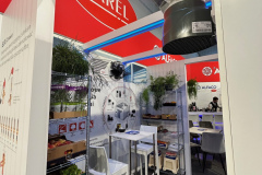 Stoisko targowe z czerwonym znakiem CAREL nad głową, białymi meblami, w tym wysokim stołem i krzesłami, plakatami informacyjnymi na ścianach, roślinami doniczkowymi i wyświetlaczami sprzętu. Przy suficie wisi duża szara jednostka wentylacyjna. projekt i nadzór Tatjana Kobuszewska trksystem.com