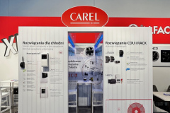 Stoisko targowe z czerwonym logo CAREL nad białymi panelami wyświetlającymi produkty sterowania HVAC, schematy techniczne i polski tekst. Broszury i urządzenia elektroniczne są zamontowane na panelach. Stołki i stoły są widoczne po lewej i prawej stronie. projekt i nadzór Tatjana Kobuszewska trksystem.com