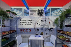 Stoisko targowe z czerwonym znakiem CAREL, stołem i dwoma białymi krzesłami oraz metalowymi półkami z warzywami i roślinami. Na tylnej ścianie znajduje się schemat rozwiązań chłodzących z polskim tekstem oraz otwór wentylacyjny nad schematem. Niebieskie światła podświetlają stoisko. projekt i nadzór Tatjana Kobuszewska trksystem.com