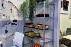 Na metalowej drucianej półce stoi zielona roślina doniczkowa, kartonowe pudełka z owocami (banany, jabłka, mandarynki), tace z broszurami i drewniane skrzynki. Obok stoją białe krzesła i stół, a na ścianie wisi duży plakat techniczny. projekt i nadzór Tatjana Kobuszewska trksystem.com