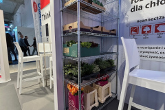 Metalowa półka z kwiatami, zielonymi roślinami, drewnianymi skrzynkami z jabłkami, pomarańczami i marchewką oraz kartonowymi pudełkami. Półka stoi na wewnętrznym stoisku eventowym z białymi krzesłami i stołami oraz banerami wyświetlającymi tekst w języku polskim w tle. projekt i nadzór Tatjana Kobuszewska trksystem.com