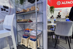 Regał z metalowego drutu mieści rośliny, pudełka, książki i skrzynki. Jest on umieszczony przy ściance działowej w nowoczesnym wnętrzu stoiska. W pobliżu znajdują się białe krzesła i stoły. Tło stanowią czerwone ściany z logo ALFACO oraz stojący i rozmawiający ludzie. projekt i nadzór Tatjana Kobuszewska trksystem.com