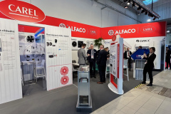 Stoisko targowe z czerwonym brandingiem CAREL i ALFACO prezentuje sprzęt chłodniczy. Kilka osób w strojach biznesowych rozmawia, podczas gdy jedna osoba robi zdjęcie smartfonem. Plakaty informacyjne i stojaki na produkty są widoczne pod jasnym oświetleniem sufitowym. projekt i nadzór Tatjana Kobuszewska trksystem.com