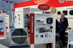 Stoisko targowe z brandingiem CAREL prezentuje sprzęt HVAC i panele informacyjne. Mężczyzna w garniturze stoi w pobliżu stoiska, gestykulując podczas rozmowy z inną osobą. Wentylatory przemysłowe i schematy produktów są widoczne na stojakach. projekt i nadzór Tatjana Kobuszewska trksystem.com