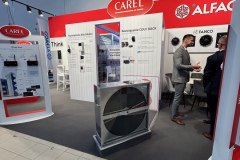 Stoisko targowe z logo CAREL i ALFA na czerwonym oznakowaniu, ekspozycje produktów na białych panelach, dwóch rozmawiających mężczyzn w garniturach, duży wentylator przemysłowy w przezroczystej obudowie na podłodze i plakaty informacyjne na temat technologii chłodzenia. projekt i nadzór Tatjana Kobuszewska trksystem.com