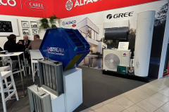 Stoisko targowe z brandingiem ALFACO i CAREL na czerwonych ścianach. Widoczne są klimatyzatory GREE i niebieski filtr powietrza XTREAM. Trzy osoby siedzą przy białym stole w tle, a podłoga jest wyłożona kafelkami i dywanem. projekt i nadzór Tatjana Kobuszewska trksystem.com