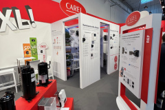 Stoisko targowe firmy CAREL zawiera białe i czerwone wyświetlacze z informacjami o produktach i urządzeniach chłodzących. Różne urządzenia elektroniczne są prezentowane na czerwonych stojakach, a plakaty wyjaśniają ich zastosowania. Przestrzeń jest dobrze oświetlona reflektorami i ma nowoczesny, industrialny sufit. projekt i nadzór Tatjana Kobuszewska trksystem.com