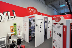 Nowoczesne stoisko wystawowe z czerwono-białym brandingiem Carel ma panele informacyjne, wyświetlacze produktów i próbki sprzętu na postumentach. Reflektory zwisają z wysokiego sufitu, a odważny napis "XL" zdobi ścianę nad małym stolikiem ze stołkami. projekt i nadzór Tatjana Kobuszewska trksystem.com