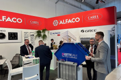 Profesjonaliści biznesowi w garniturach rozmawiają na stoisku targowym z czerwonym oznakowaniem ALFACO i Carel nad głową. Stoły ekspozycyjne, krzesła i niebieska maszyna przemysłowa z napisem "XSTREAM". Stoiska ALFACO i GREE są widoczne w nowoczesnej, dobrze oświetlonej przestrzeni. projekt i nadzór Tatjana Kobuszewska trksystem.com