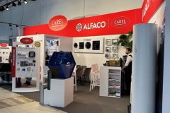 Stoisko wystawiennicze ALFACO i CAREL z czerwono-białym brandingiem. Stoisko prezentuje sprzęt HVAC, broszury i plakaty produktów. Dwa krzesła i stół są ustawione wewnątrz; osoba stoi w pobliżu monitora i materiałów promocyjnych. projekt i nadzór Tatjana Kobuszewska trksystem.com