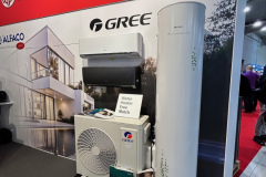 Stoisko targowe z logo ALFACO i CAREL na czerwonej ścianie. Prezentowane są urządzenia GREE HVAC: klimatyzator ścienny, jednostka zewnętrzna i wysoki biały podgrzewacz wody. Napis głosi Water Heater Heat Pump Match. W tle widoczni są ludzie. projekt i nadzór Tatjana Kobuszewska trksystem.com