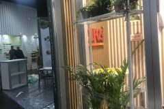 Stoisko targowe z drewnianymi listwami, półkami z zielonymi roślinami doniczkowymi i czerwonym napisem DOLFOS. W tle ludzie ustawiają kolejne stoisko ze sprzętem i roślinami na podłodze. projekt i nadzór Tatjana Kobuszewska trksystem.com