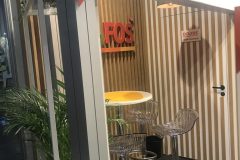 Nowoczesne stoisko wystawowe z drewnianymi ścianami, przezroczystymi plastikowymi stołkami barowymi, małym okrągłym stolikiem, zielonymi roślinami i napisem "DOLFOS PETS - zasady prawidłowego żywienia". Jasne oświetlenie podkreśla przytulną część wypoczynkową. projekt i nadzór Tatjana Kobuszewska trksystem.com