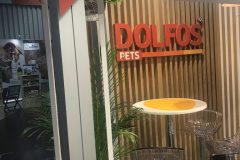 Stoisko DOLFOS PETS z drewnianą ścianą z listew, czerwonym logo, okrągłym biało-żółtym stołem i przezroczystymi stołkami barowymi. W tle widoczna jest zielona roślina i inne stoisko. projekt i nadzór Tatjana Kobuszewska trksystem.com