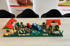 Ekspozycja stołowa prezentuje miniaturową scenę farmy jako Stoisko reklamowe Dolfos, zawierającą zabawkowe budynki z napisem DOLFOS, zwierzęta, pojazdy, ogrodzenia i drzewa ustawione na jasnym drewnianym stole z czarnymi krzesłami w tle. projekt i nadzór Tatjana Kobuszewska trksystem.com