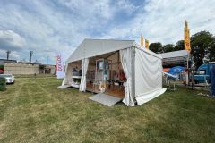 Biały namiot eventowy stoi na trawiastym polu pod częściowo zachmurzonym niebem. Namiot jest otwarty, a w środku znajdują się stoły, krzesła i materiały Stoisko reklamowe Dolfos. Namiot otaczają flagi i banery, a w tle widać drzewa i budynki. projekt i nadzór Tatjana Kobuszewska trksystem.com