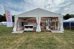 Biały namiot wystawowy z otwartymi bokami służy jako Stoisko reklamowe Dolfos, w którym znajdują się broszury, wyświetlacze, obrazy i lada. Ustawiony na trawie z flagą i innymi namiotami w pobliżu, stoi pod częściowo zachmurzonym niebem. projekt i nadzór Tatjana Kobuszewska trksystem.com