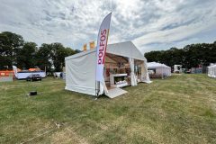 Biały namiot festiwalowy z flagą POLFOS stoi na trawiastym polu pod zachmurzonym niebem. W tle widoczne są inne namioty, flagi i drzewa. Wewnątrz namiotu znajduje się Stoisko reklamowe Dolfos ze stołami i wystawami. projekt i nadzór Tatjana Kobuszewska trksystem.com