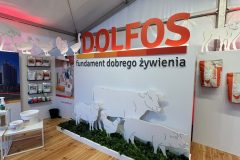 Stoisko reklamowe Dolfos zawiera duży czerwony tekst, białe wycinanki zwierząt gospodarskich na ścianie z napisem "fundament dobrego żywienia", drewnianą podłogę i półki z produktami - tworząc zachęcającą i informacyjną ekspozycję. projekt i nadzór Tatjana Kobuszewska trksystem.com