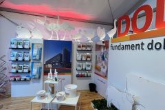 Stoisko targowe z drewnianą podłogą prezentuje produkty na półkach i stołach. Stoisko reklamowe Dolfos przedstawia krowy i inne zwierzęta hodowlane, zieleń, opakowania produktów i duży napis DOL na ścianie. projekt i nadzór Tatjana Kobuszewska trksystem.com