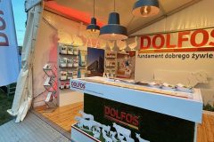 Stoisko reklamowe Dolfos zawiera sylwetki zwierząt, broszury i lampy wiszące wewnątrz białego namiotu. Stoisko obejmuje trawiastą ladę i wyświetla materiały promocyjne na półkach, aby przyciągnąć odwiedzających. projekt i nadzór Tatjana Kobuszewska trksystem.com