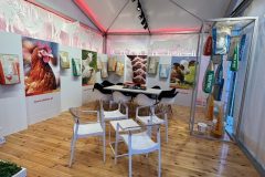 Stoisko reklamowe Dolfos ma drewnianą podłogę, białe krzesła i stół. Kolorowo zapakowane produkty, plakaty kurczaków i zwierząt hodowlanych wzdłuż ścian, podkreślone reflektorami i szklaną gablotą po prawej stronie. projekt i nadzór Tatjana Kobuszewska trksystem.com