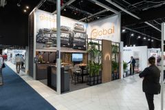 Stoisko targowe Global Motion w Ptak Warsaw Expo zawiera obrazy pojazdów, oznakowanie marki, strefę do siedzenia i zieloną roślinność, a ludzie spacerują i wchodzą w interakcje wokół tętniącej życiem przestrzeni wystawienniczej. projekt i nadzór Tatjana Kobuszewska trksystem.com