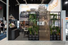 Stoisko targowe Global Motion dla Global Group w Ptak Warsaw Expo zawiera odważny branding, zdjęcia ciężarówek, część wypoczynkową, czarną ladę, rośliny doniczkowe i drewnianą ściankę działową, a w tle przechadzają się ludzie. projekt i nadzór Tatjana Kobuszewska trksystem.com