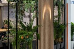 Nowoczesna przegroda wewnętrzna z czarnymi metalowymi ramami, zielonymi roślinami doniczkowymi i pionowym panelem z drewnianych listew definiuje to Stoisko targowe Global Motion w Ptak Warsaw Expo w stylowej przestrzeni komercyjnej. projekt i nadzór Tatjana Kobuszewska trksystem.com