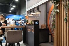 Nowoczesne Stoisko targowe Global Motion w Ptak Warsaw Expo obejmuje elegancką czarną ladę, stołki, bujne rośliny i uderzającą dużą grafikę pojazdu na ścianie, z ludźmi mieszającymi się w tle. projekt i nadzór Tatjana Kobuszewska trksystem.com