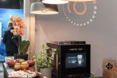 Nowoczesna stacja kawowa w Stoisku targowym Global Motion, Ptak Warsaw Expo, z samoobsługowym ekspresem do kawy, talerzem jabłek, doniczkową orchideą i lampami wiszącymi nad głową. projekt i nadzór Tatjana Kobuszewska trksystem.com