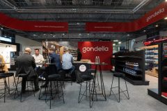 Czterech mężczyzn siedzi przy wysokich stołach i dyskutuje w stoisku targowym Gondella podczas targów Targi Retail Show, otoczonych regałami i materiałami promocyjnymi w Hali EXPO XXI. projekt i nadzór Tatjana Kobuszewska trksystem.com