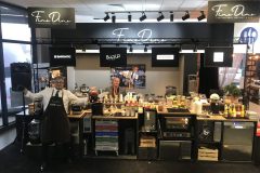 Dwóch mężczyzn w fartuchach stoi za ladą kawiarni Hendi Sweet słodkie stoisko, otoczone owocami, słoikami i sprzętem, pod szyldem Fine Dine podczas imprezy pod dachem. projekt i nadzór Tatjana Kobuszewska trksystem.com