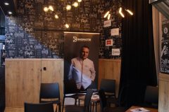 Mała część wypoczynkowa z czarnymi krzesłami, drewnianą podłogą i banerem przedstawiającym szefa kuchni pod nowoczesnym oświetleniem wiszącym w stoisku targowym Hendi w Fine Dine, ze ścianą w stylu czarnej tablicy w tle. projekt i nadzór Tatjana Kobuszewska trksystem.com