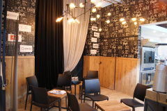 Mała kawiarnia na stoisku targowym.  z czarnymi krzesłami, drewnianymi stołami, dekoracyjnym oświetleniem i tapetą w stylu tablicy z motywami kawy i logo Fine Dine. warsawsweettech.com  . projekt i nadzór Tatjana Kobuszewska trksystem.com