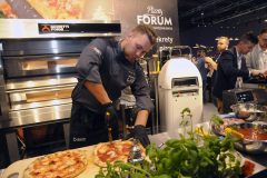 Kucharz w czarnym uniformie kroi pizzę w Expo XXI podczas Forum Szefów Kuchni i Restauratorów, z piecami Hendi, świeżymi składnikami i obserwatorami w tle. projekt i nadzór Tatjana Kobuszewska trksystem.com