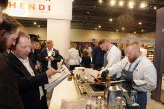 Kilku szefów kuchni przygotowuje jedzenie na stoisku, podczas gdy uczestnicy obserwują, czytają broszury i gromadzą się na Forum Szefów Kuchni i Restauratorów podczas targów Expo XXI. projekt i nadzór Tatjana Kobuszewska trksystem.com